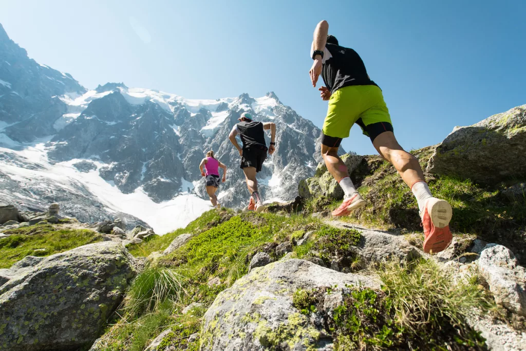 Three trail runners, two men and a woman, running up a steep trail in the mountains in the Alps on a hot, bright summer day.